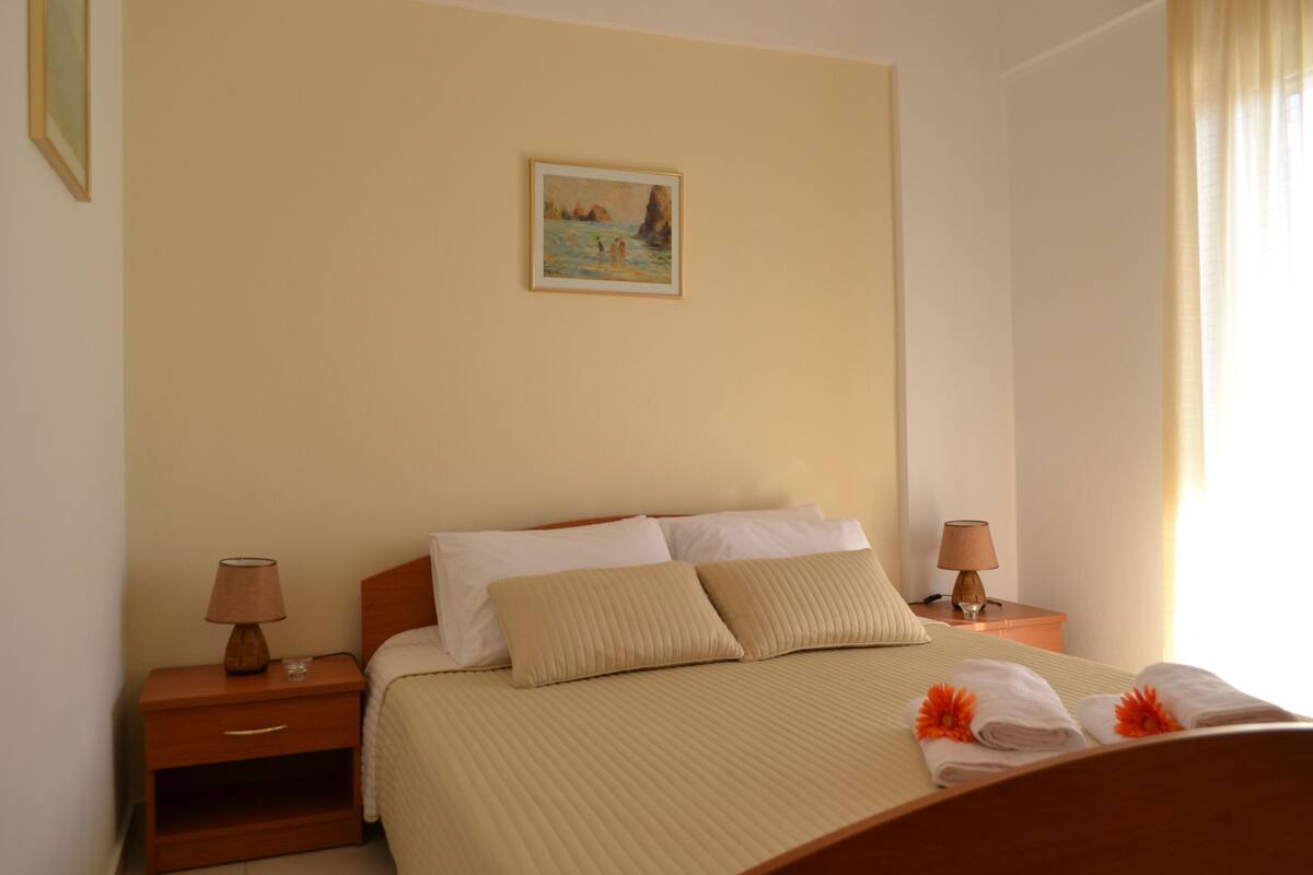 A neatly made bed with a beige bedspread and pillows, two bedside tables with lamps, a painting on the wall, and a window with curtains.