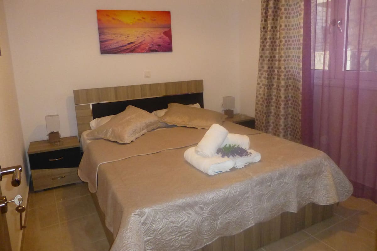 A hotel room with a bed, a nightstand, a lamp, a painting on the wall, and a patterned curtain.