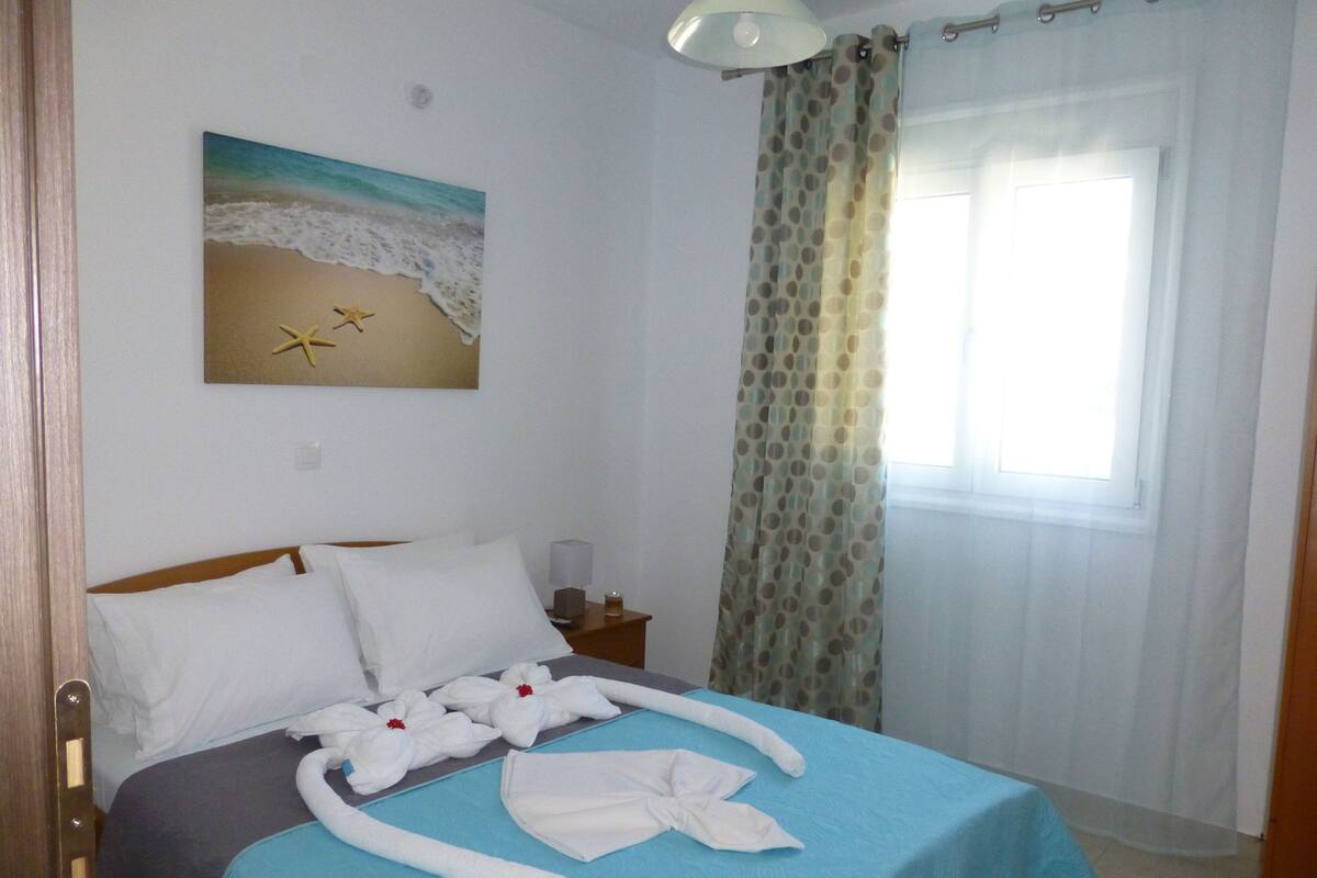 A hotel room with a bed, a painting of a beach, a window with curtains, and a nightstand with a lamp.