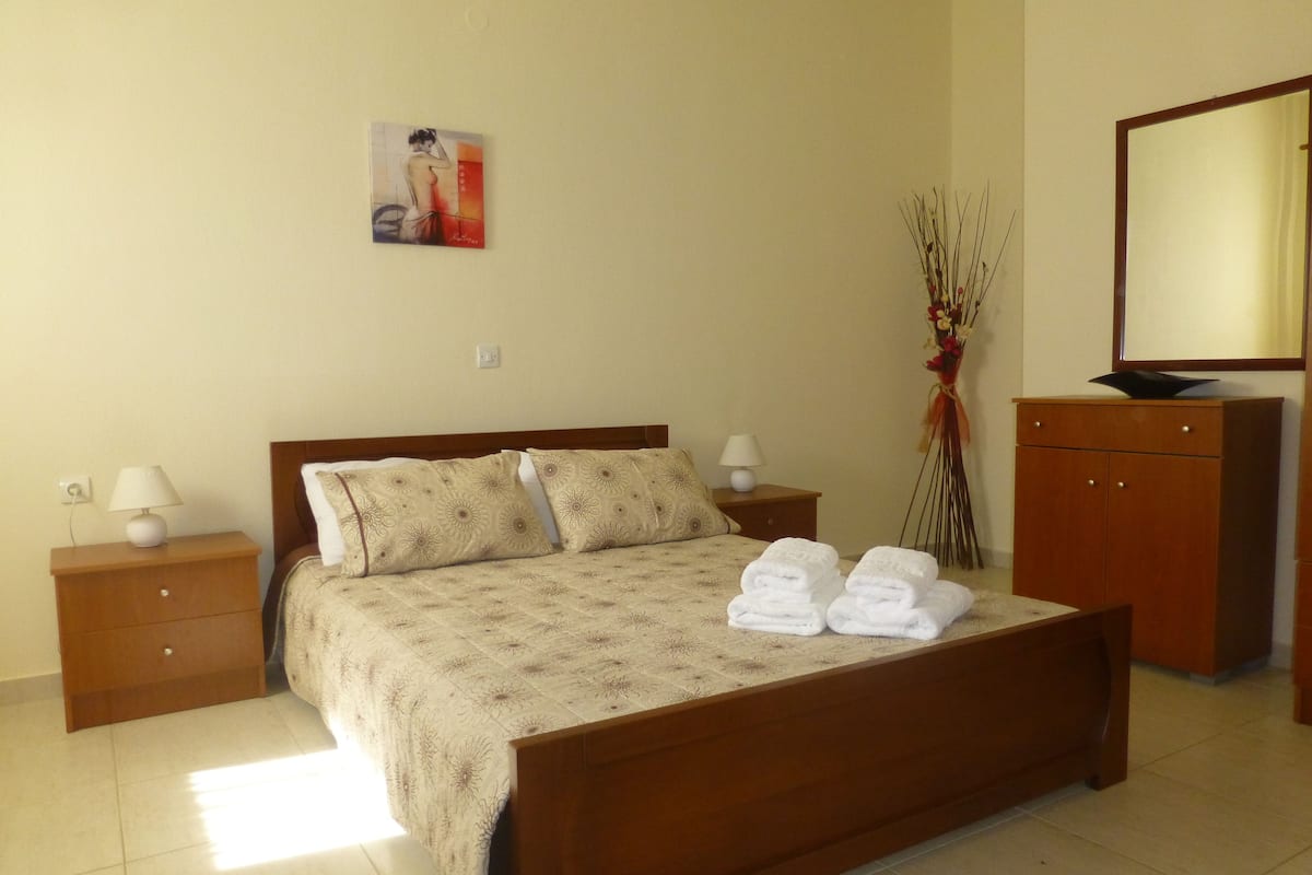 A neatly made bed with a floral-patterned bedspread, two pillows, and two folded towels on the bed. There is a wooden nightstand with a lamp on each side of the bed, a wooden dresser with a mirror, and a wall-mounted painting above the bed.