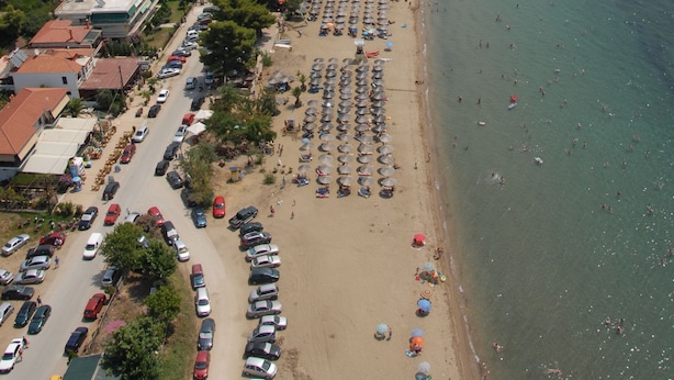 An aerial view of a coastal town with a beach, parked cars, and buildings with red roofs. - Pavloudis Apartments (Polygyros)