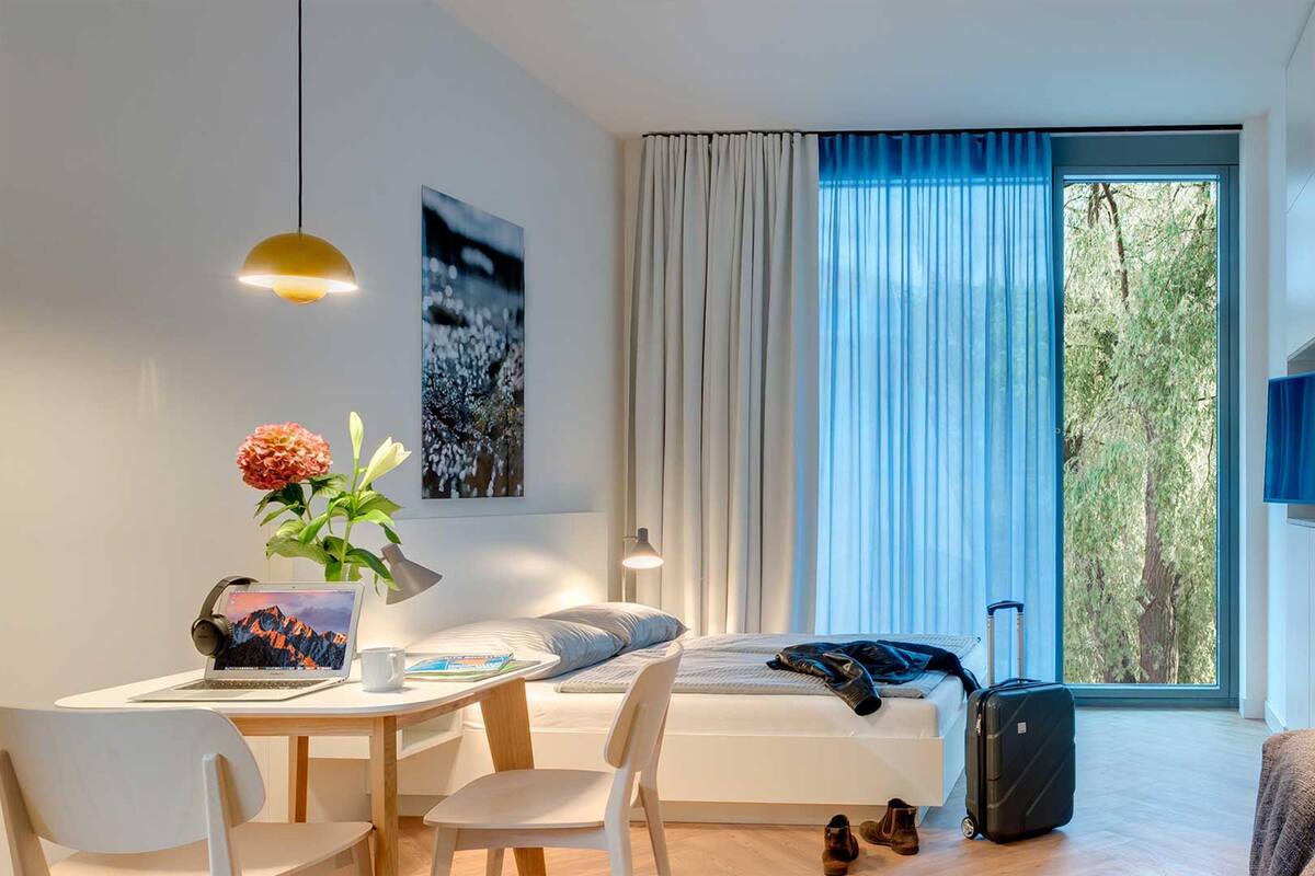 A modern hotel room with a bed, a desk with a laptop, a chair, a table with a vase of flowers, and a suitcase.