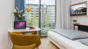A modern bedroom with a bed, a desk with a laptop, a chair, and a large window with curtains.