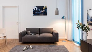 A modern living room with a grey sofa, a laptop, and a pair of shoes on a rug. There is a picture on the wall, a floor lamp, and a plant on a table. - smartments Berlin City-West (Berlin)