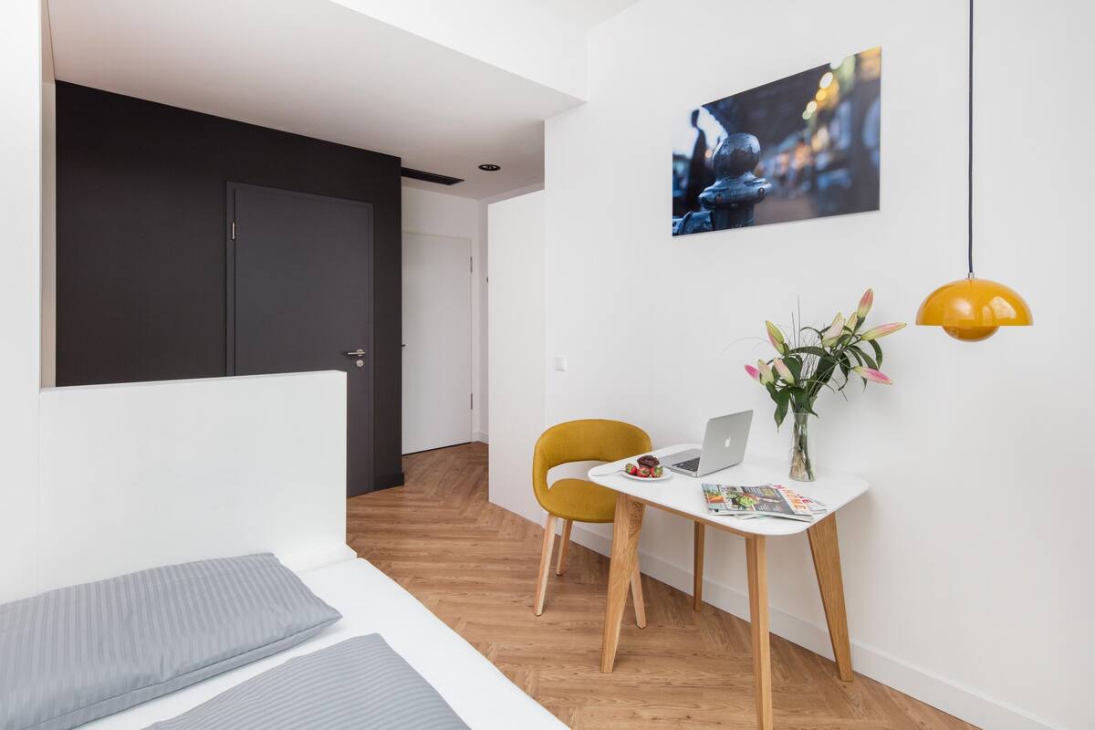 A modern room with a bed, a desk with a laptop, a chair, a vase with flowers, and a painting on the wall.