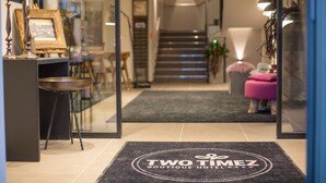 Zona de recepción de hotel con felpudo, escalera y área de descanso con un otoman rosa.