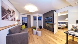 Una sala de estar moderna con un sofá, una mesa de centro y un televisor. Se observa una cama al fondo y una ventana con cortinas.
