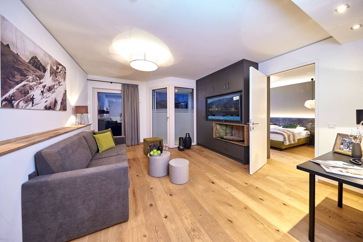 Een moderne woonkamer met een bank, een salontafel en een televisie. Achterin staat een bed en er is een raam met gordijnen.