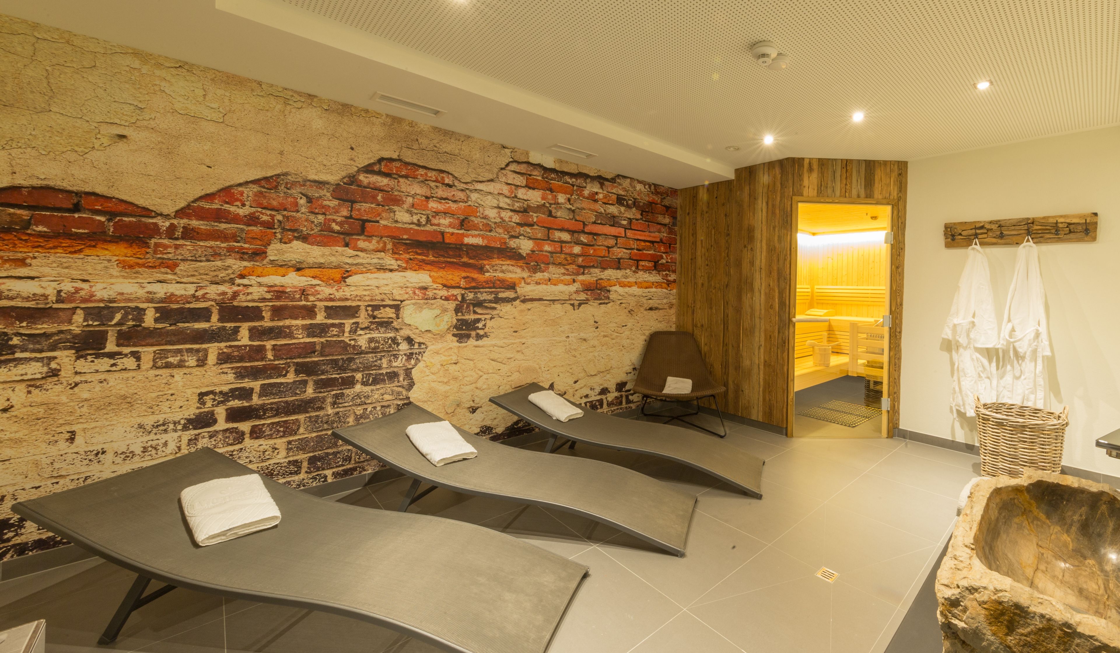 Un cuarto de spa con cuatro sillones reclinables, una pared de ladrillo, paneles de madera, un lavabo de piedra y una puerta de madera que da a una sauna.