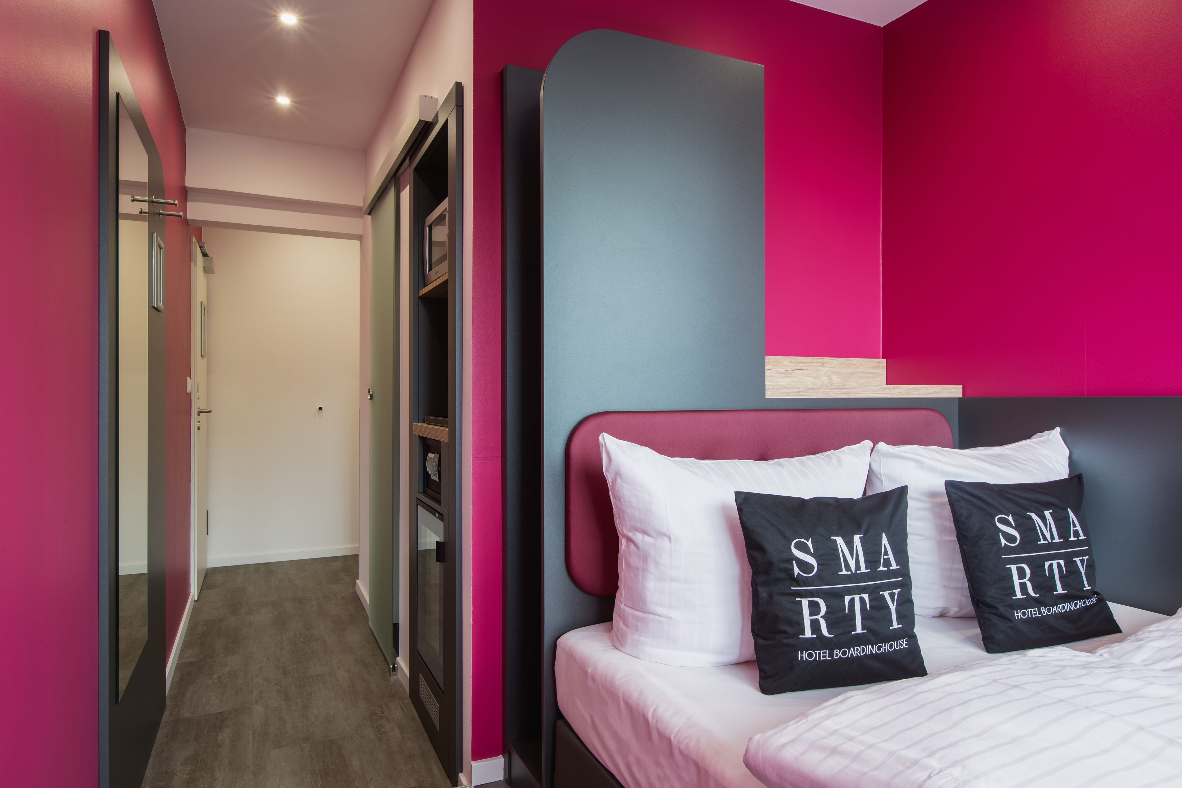 Une chambre d’hôtel moderne aux couleurs rose et gris, avec un lit doté d’oreillers blancs et noirs, et une armoire avec un miroir.