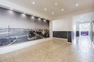 Um corredor amplo com um grande mural de paisagem urbana na parede, piso de azulejos e um banco ao fundo.