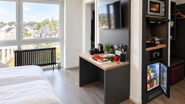 A modern hotel room with a bed, a small kitchenette, a TV, and a view of the city.