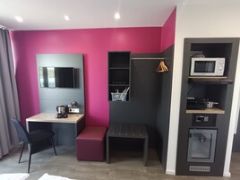 Quarto compacto com escrivaninha, um banco, TV fixada na parede, micro-ondas e um armário pequeno.