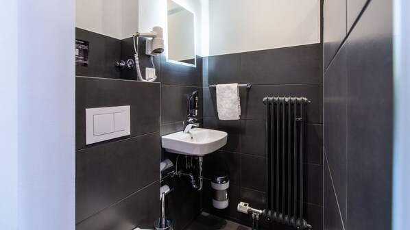 Un baño moderno con un lavamanos blanco, baldosas oscuras, un perchero, un secador de pelo y un radiador.