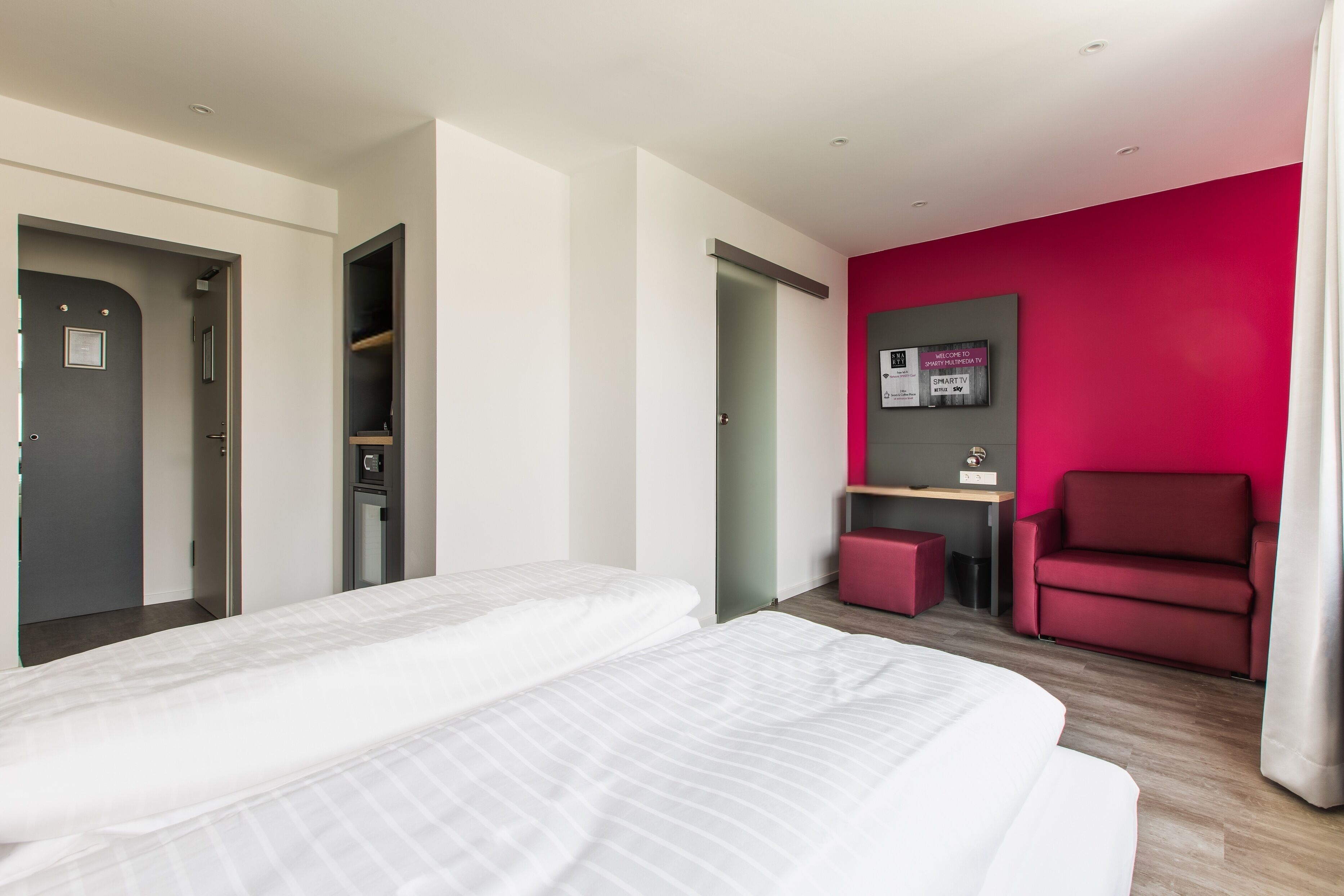 A modern hotel room with a bed, a desk, a red chair, and a bright pink accent wall.