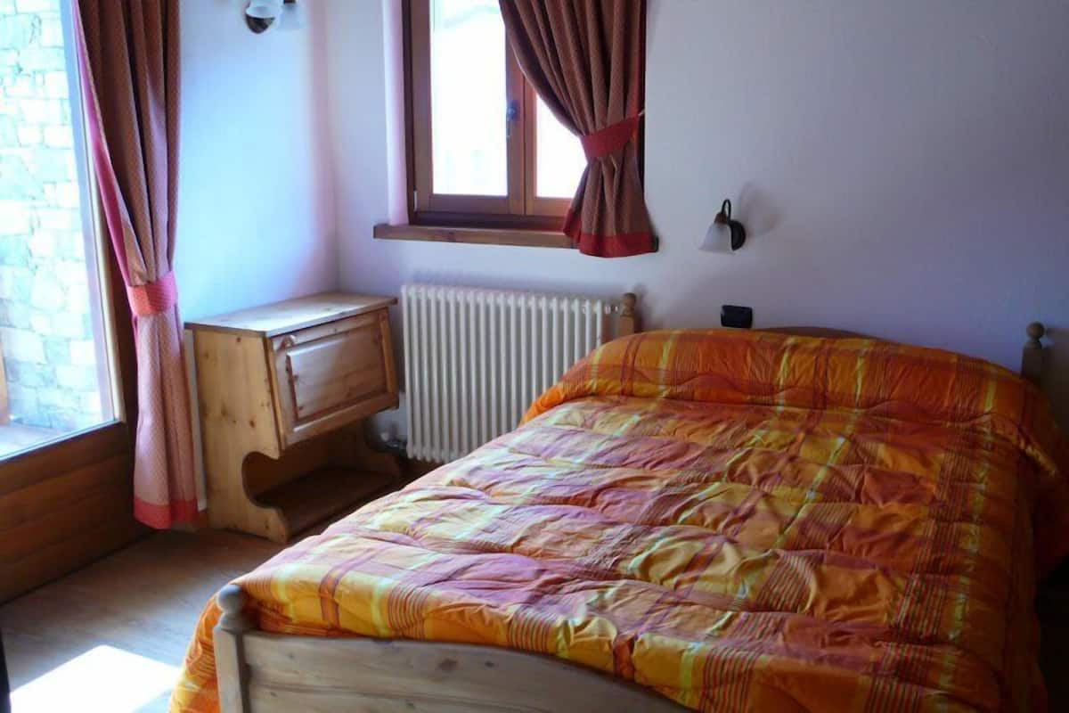 A bedroom with a wooden bed, a nightstand, a window with curtains, and a radiator.