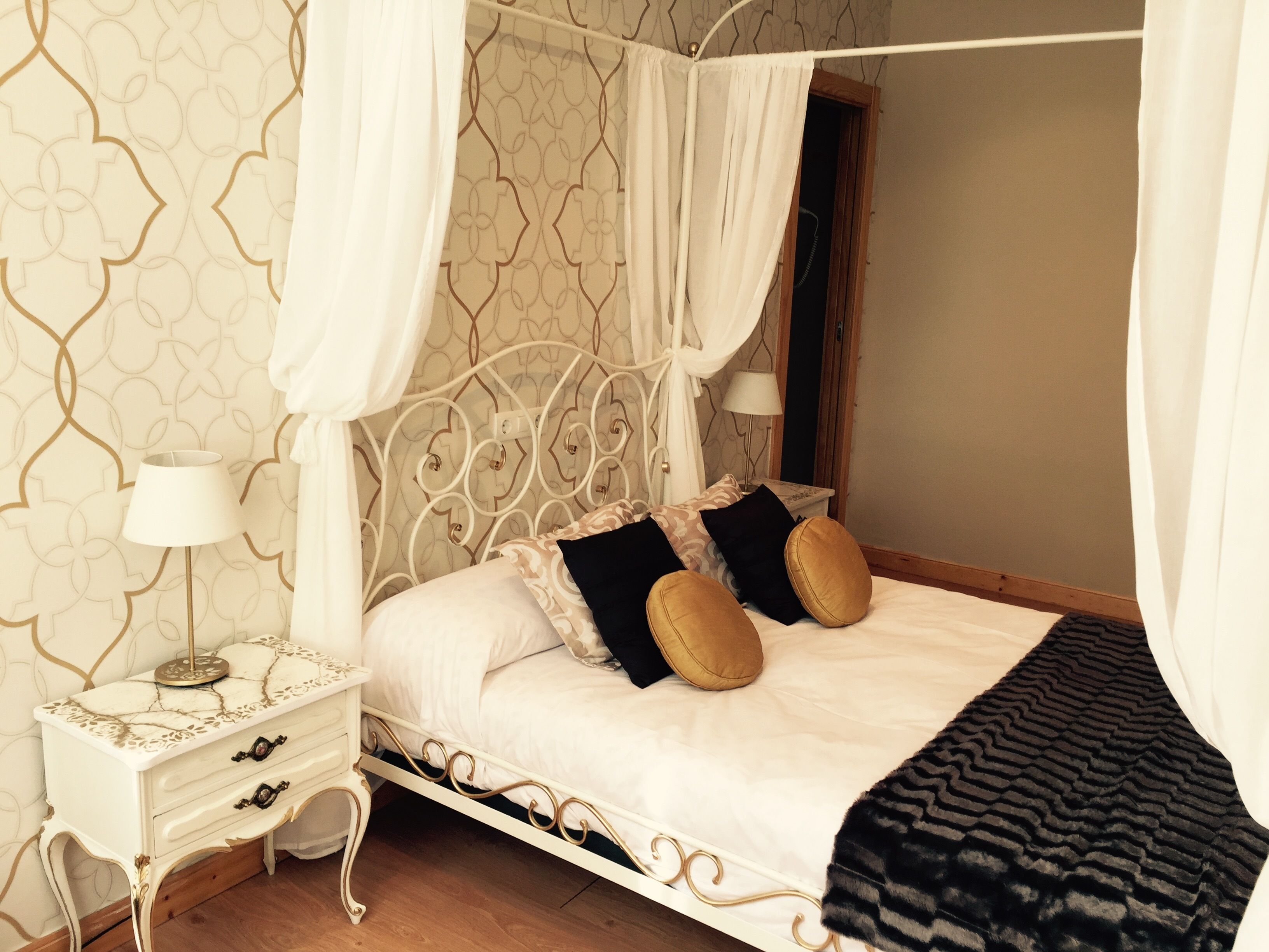 Ein Himmelbett mit goldfarbenem Metallgestell, ein Nachttisch mit Lampe und Wandpapier mit Muster.