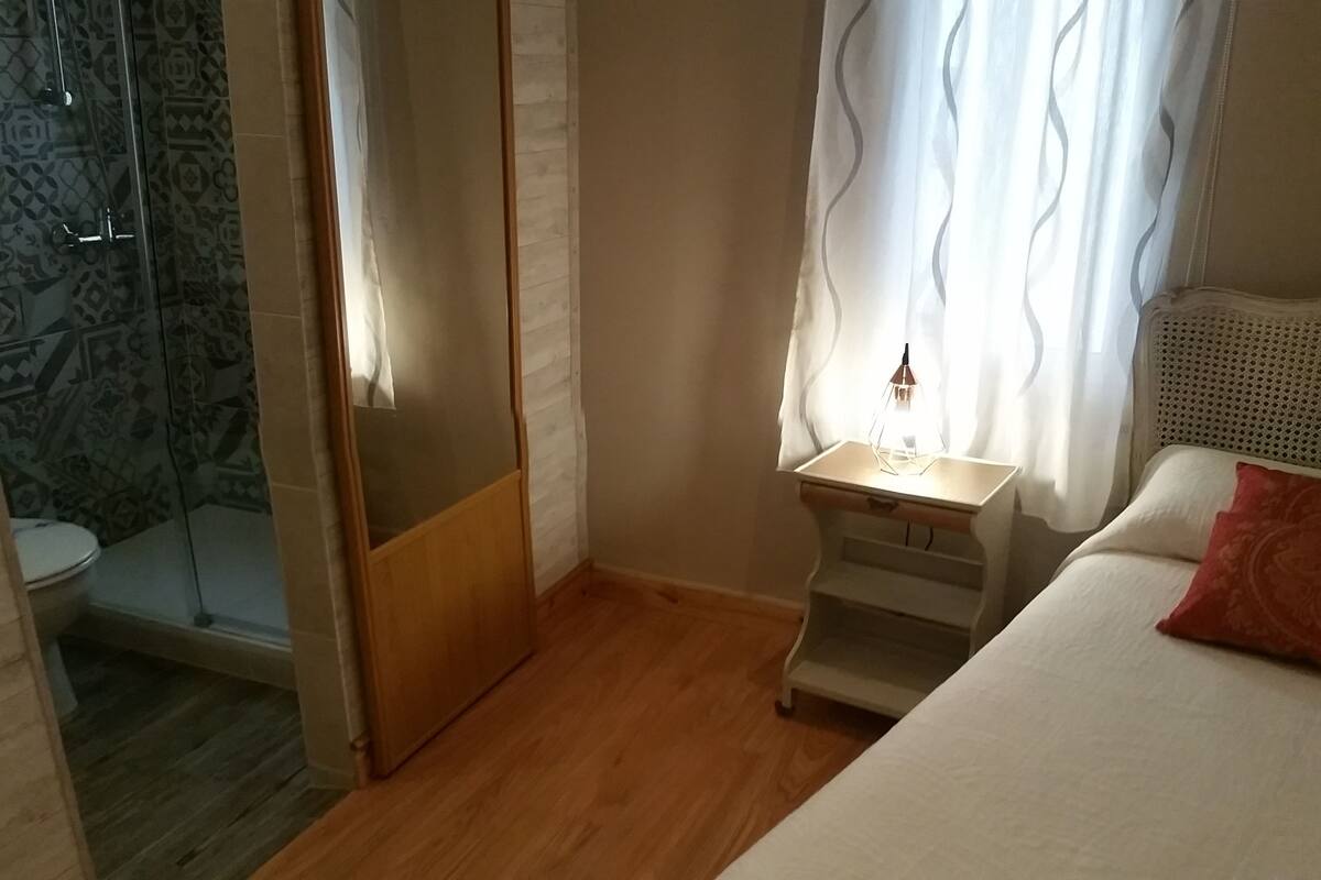 Une chambre à coucher avec un lit, une table de chevet avec une lampe, une fenêtre avec des rideaux et un espace douche avec un mur décoré de motifs.