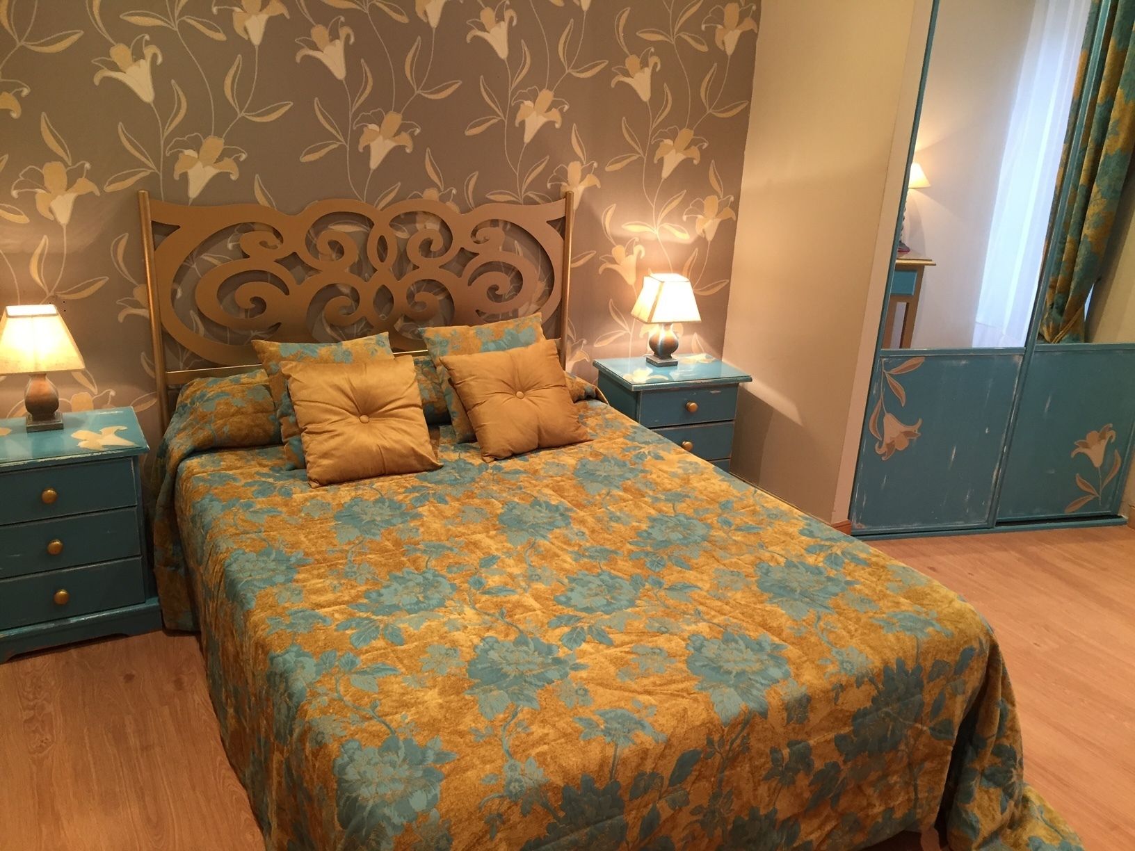 Un dormitorio con una colcha de estampado floral, dos mesitas de noche con lámparas y un cabecero decorativo.