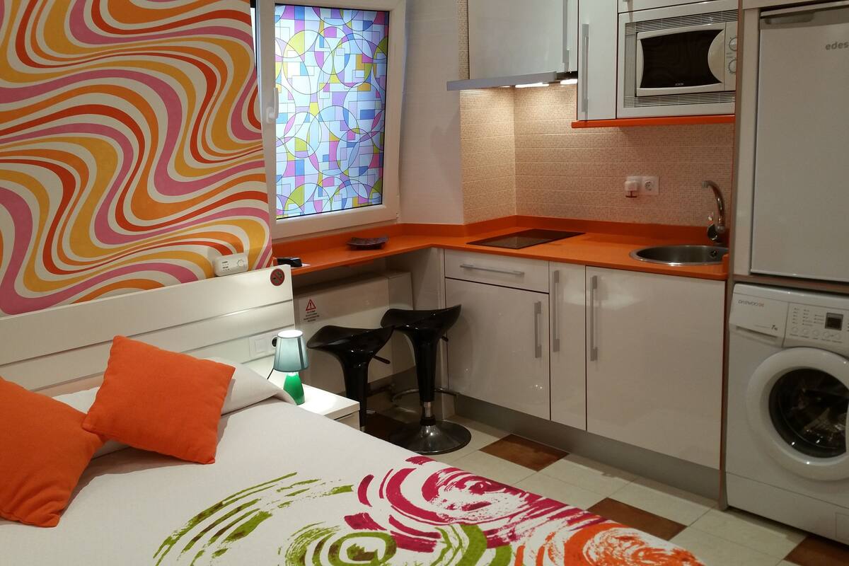 Une chambre d’hôtel compacte comprenant un lit, une petite kitchenette, un lave-linge et une fenêtre ornée d’un motif abstrait aux couleurs vives.