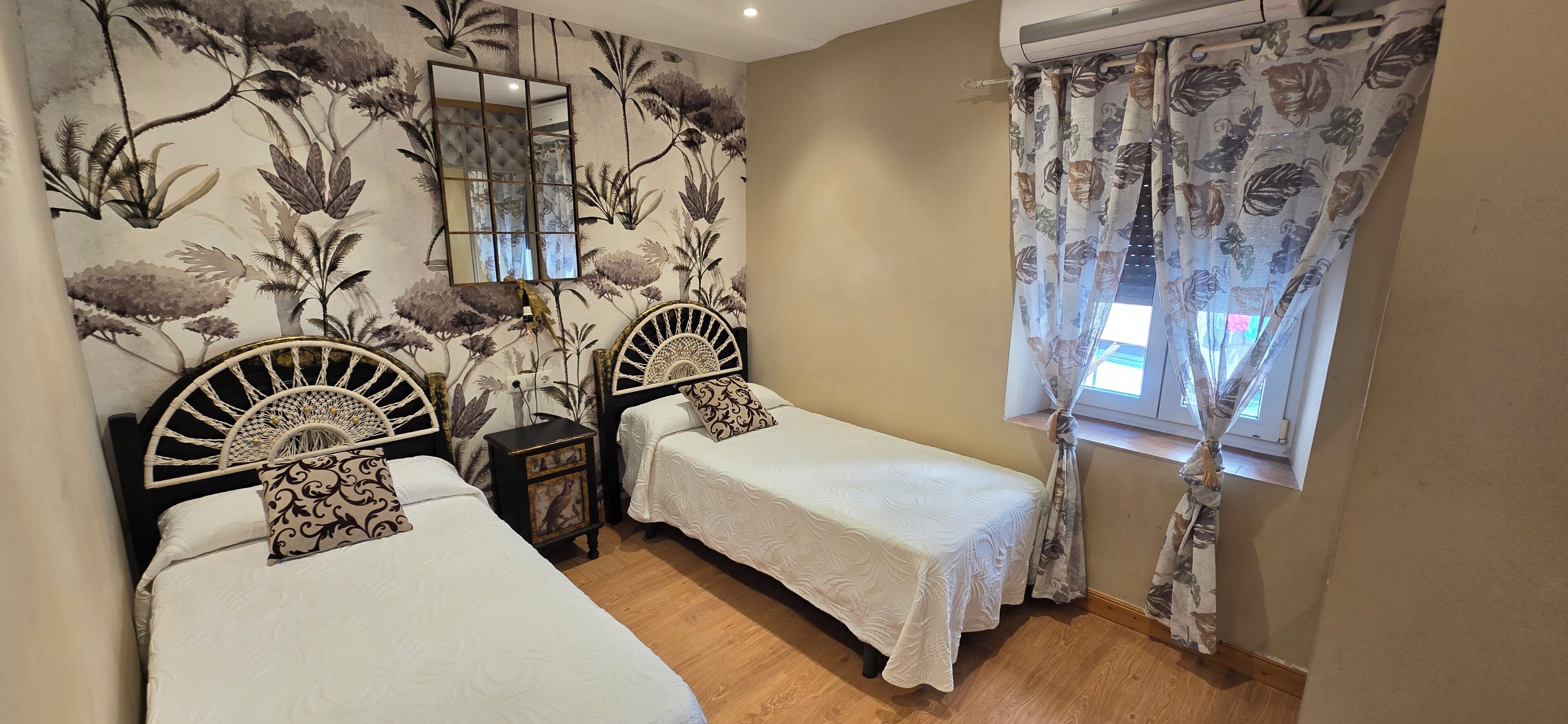 A bedroom with two single beds, a nightstand with a lamp, a mirror, and a window with floral curtains.