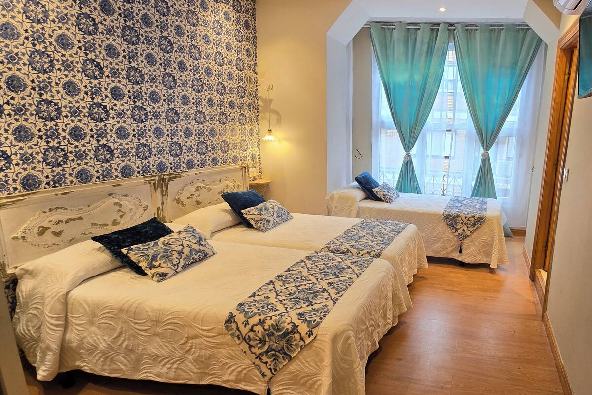 Une chambre d’hôtel avec deux lits, une télévision et une vue sur un immeuble, visible à travers une fenêtre ornée de rideaux verts.