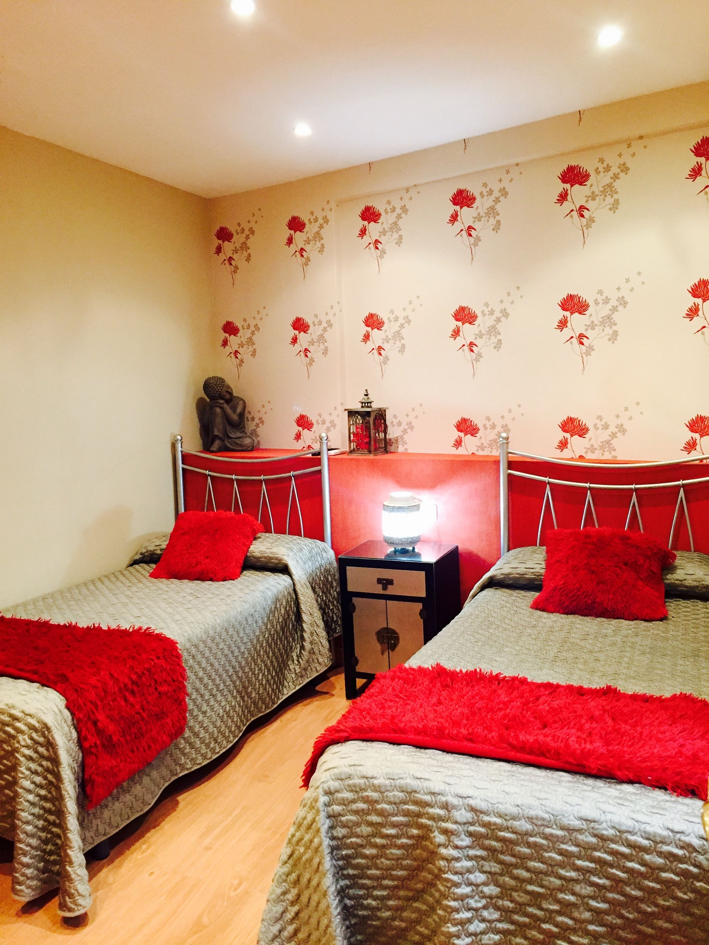 Una habitación con dos camas individuales, cada una con una manta roja, una mesita con lámpara y una pared cubierta de papel tapiz floral.