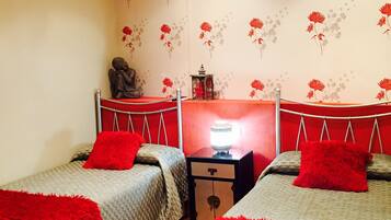 Una habitación con dos camas individuales, cada una con una manta roja, una mesita con lámpara y una pared cubierta de papel tapiz floral.