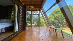 Um deck de madeira com uma cama e uma mesa, janelas amplas com vista para árvores.