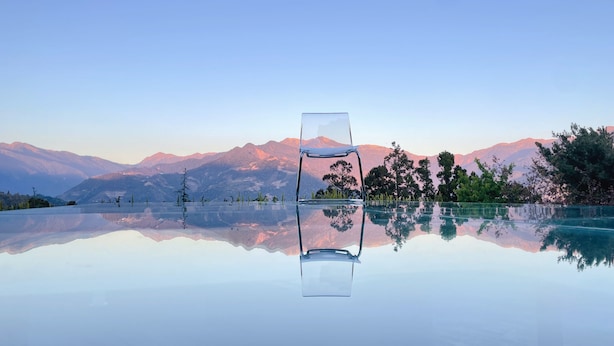 Mesa de vidro transparente com uma cadeira no centro, refletindo uma paisagem montanhosa.