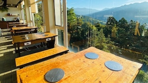 Uma área de refeições com mesas e bancos de madeira, grandes janelas com vista para montanhas e vegetação.