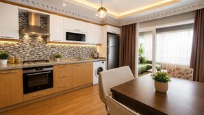 A modern kitchen with a built-in oven, microwave, and refrigerator, a dining area with a table and chairs, and a view of a green sofa through a glass door. - Real King Residence  (Yomra)