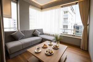 Un salón moderno con un sofá gris, una mesa de centro de madera con pasteles, y vistas a edificios a través de grandes ventanales.