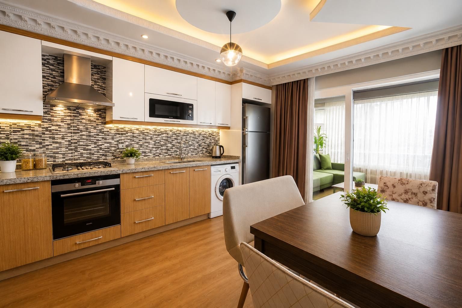 Una cocina moderna con horno empotrado, microondas y refrigerador, un comedor con mesa y sillas, y vista a un sofá verde a través de una puerta de vidrio.