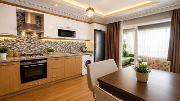 Una cocina moderna con horno empotrado, microondas y refrigerador, un comedor con mesa y sillas, y vista a un sofá verde a través de una puerta de vidrio.