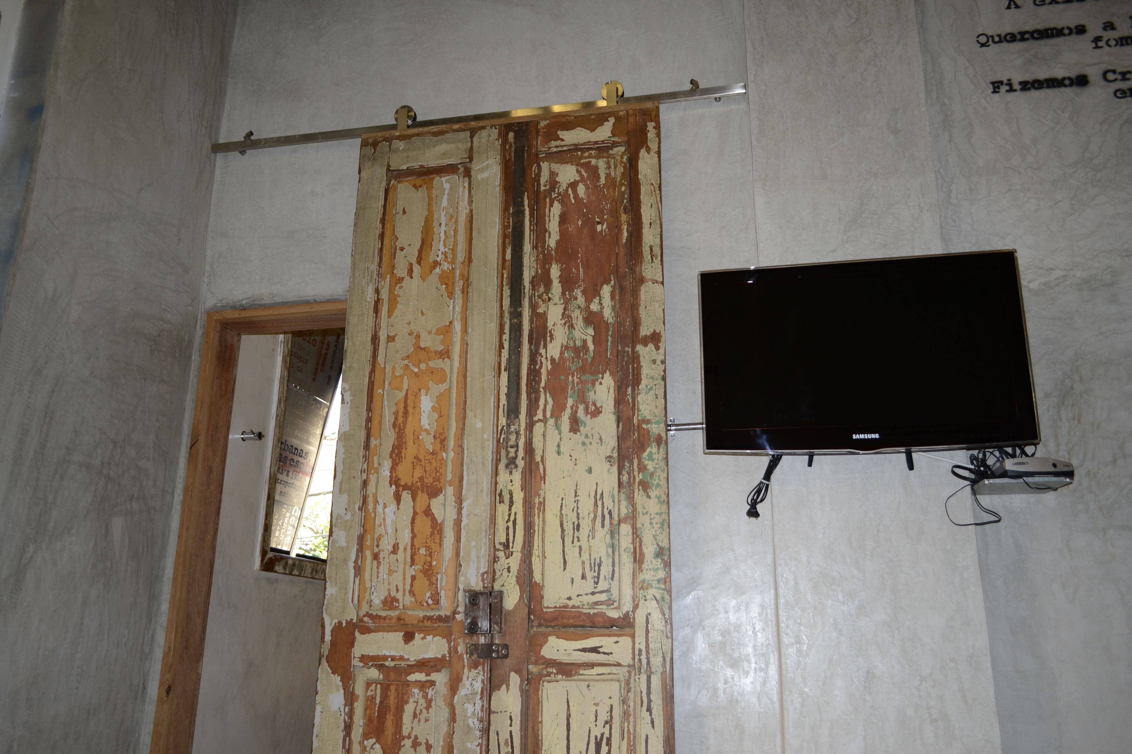 Una habitación con un televisor montado en la pared, una puerta con pintura descascarada y una ventana.