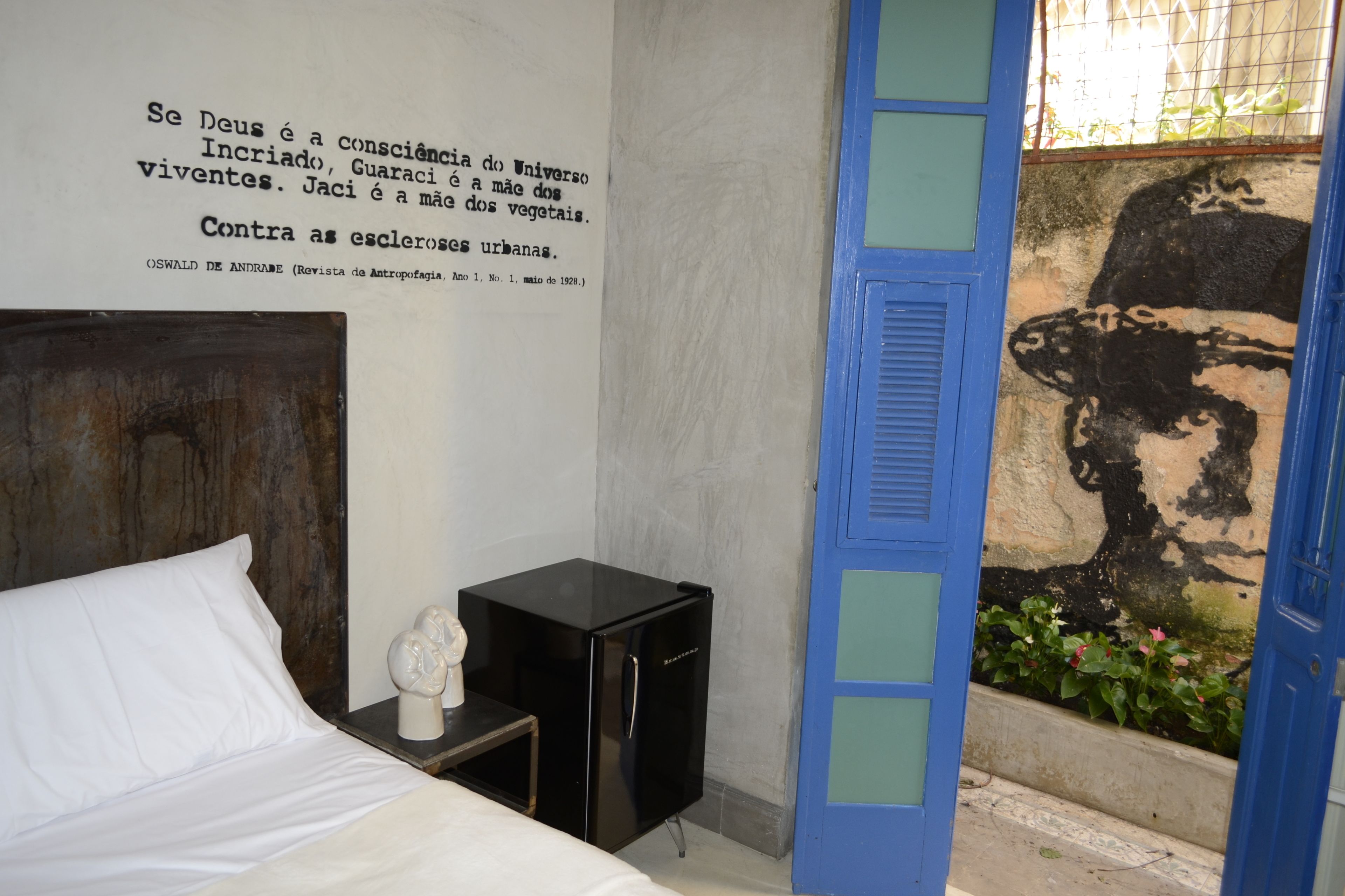 A bedroom with a bed, a small black cabinet, a white bust sculpture, and a wall with a black and white mural.