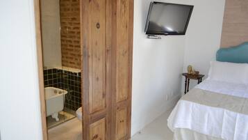 Un dormitorio con un armario de madera, un televisor montado en la pared, una cama con cubrecama blanco y beige, y una mesita de noche con un florero.