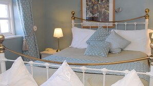 Una cama con estructura blanca y detalles dorados, una mesita de noche con lámpara, y un cuadro en la pared.