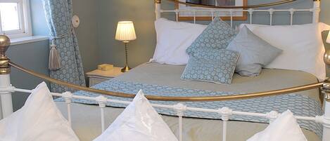 Una cama con estructura blanca y detalles dorados, una mesita de noche con lámpara, y un cuadro en la pared.