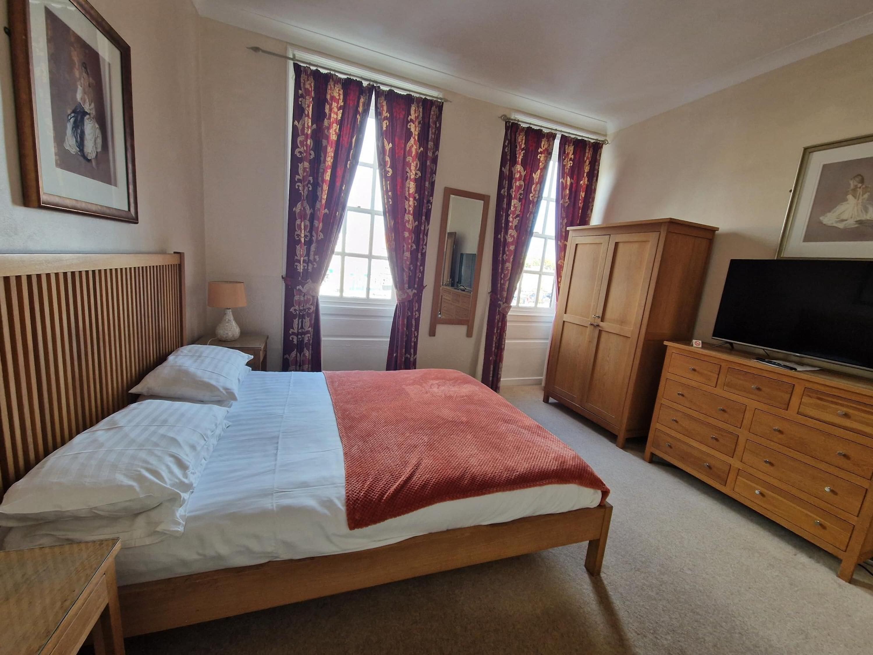 A bedroom with a bed, wooden headboard, a television on a wooden cabinet, and two windows with curtains.