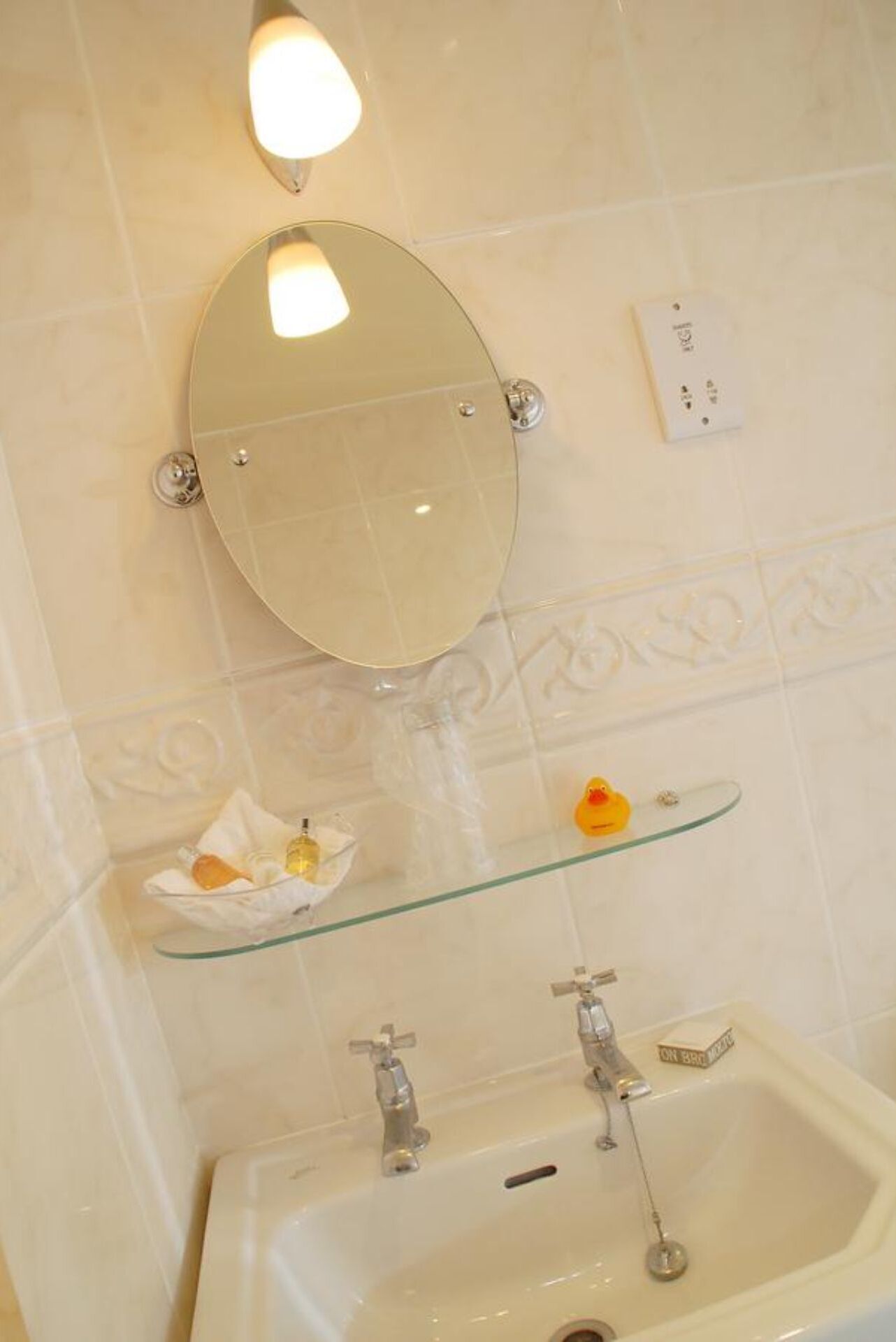 A bathroom with a mirror, a glass shelf with toiletries, and a sink with faucets.