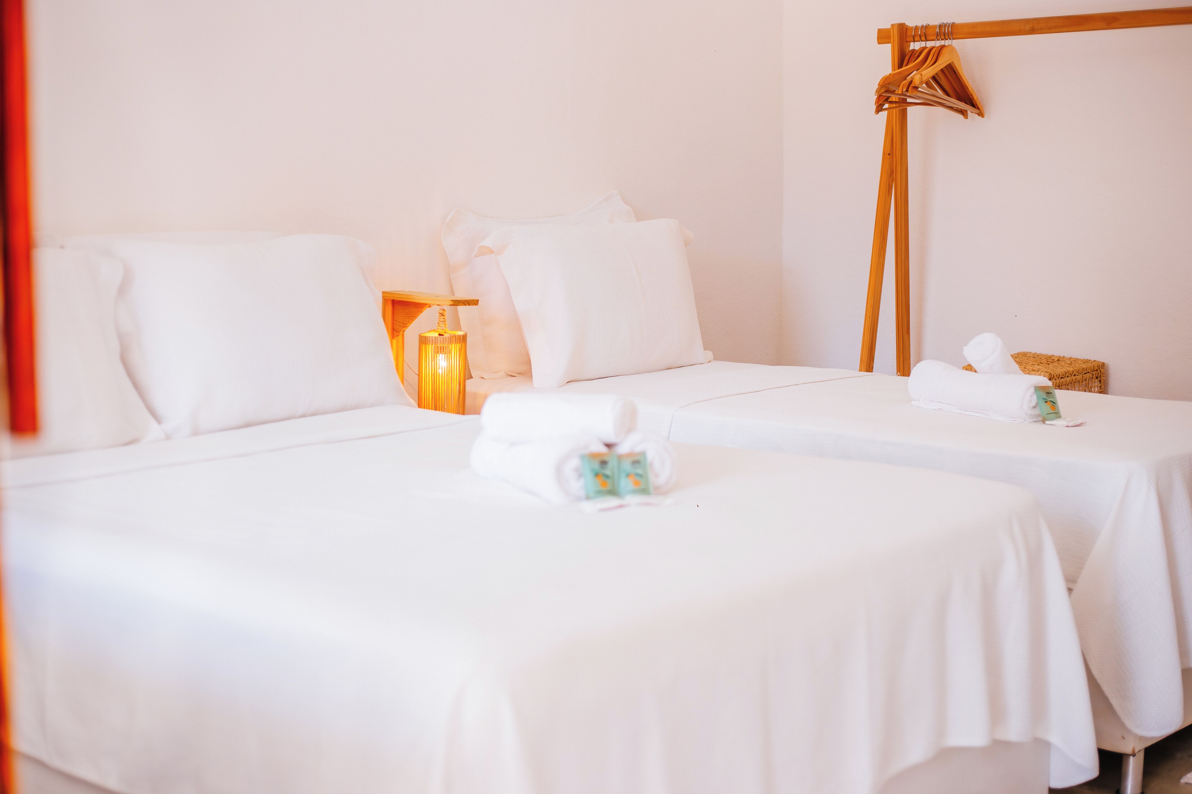 Habitación de hotel con dos camas, ropa de cama blanca, una mesita de noche con lámpara y un tozalengua de madera.