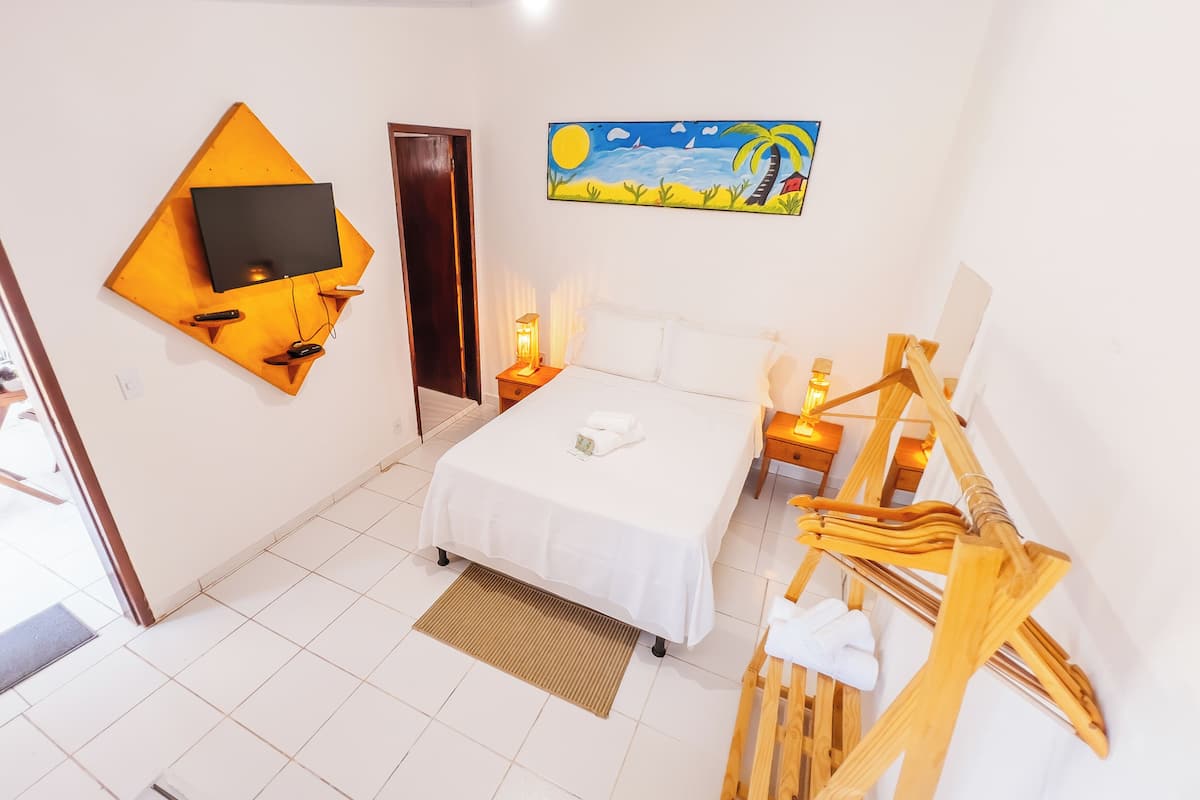 A hotel room with a bed, a TV mounted on the wall, a painting of a beach scene, and a wooden chair.