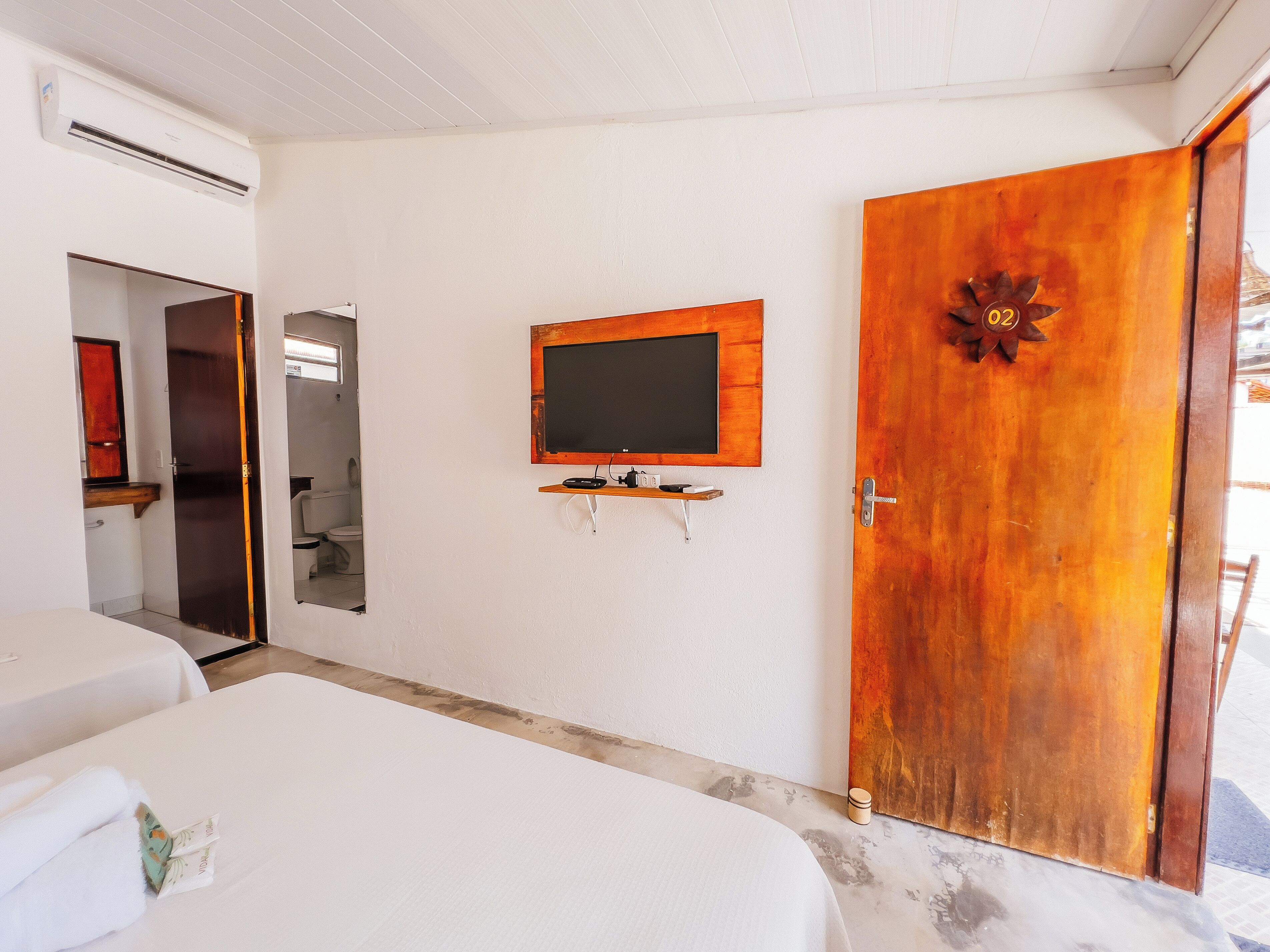 Habitación de hotel con puerta de madera, televisor montado, estante con objetos y un espejo.