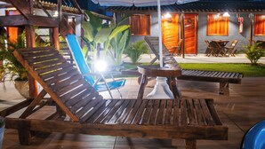 A wooden lounge chair and table under a striped umbrella, with a wooden building in the background. - Pousada do Enildo (São Miguel dos Milagres)