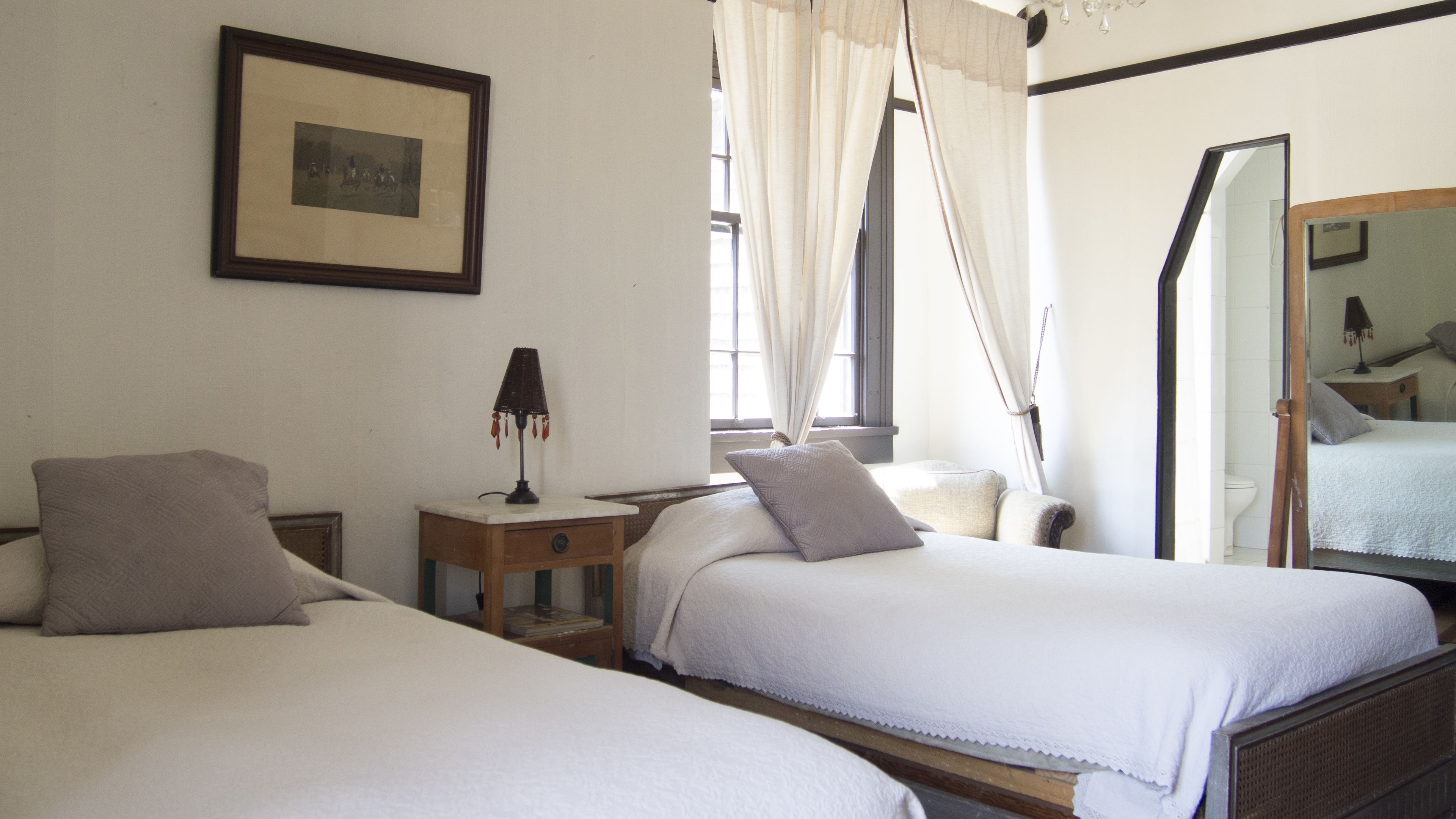 Une chambre avec deux lits, une table de chevet avec une lampe, un tableau encadré au mur et une fenêtre avec des rideaux.