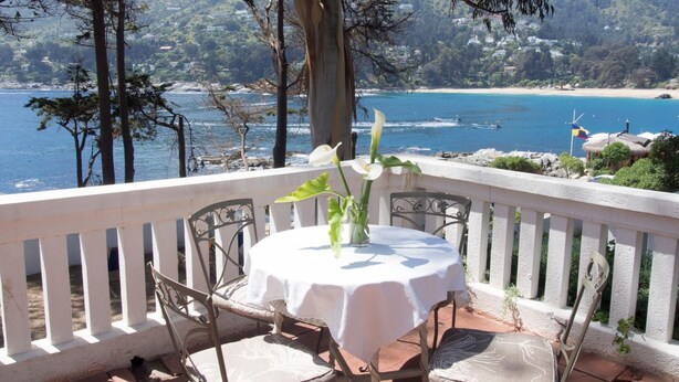 Un balcón con mesa y sillas, con vistas a un paisaje costero que incluye una playa y el océano.