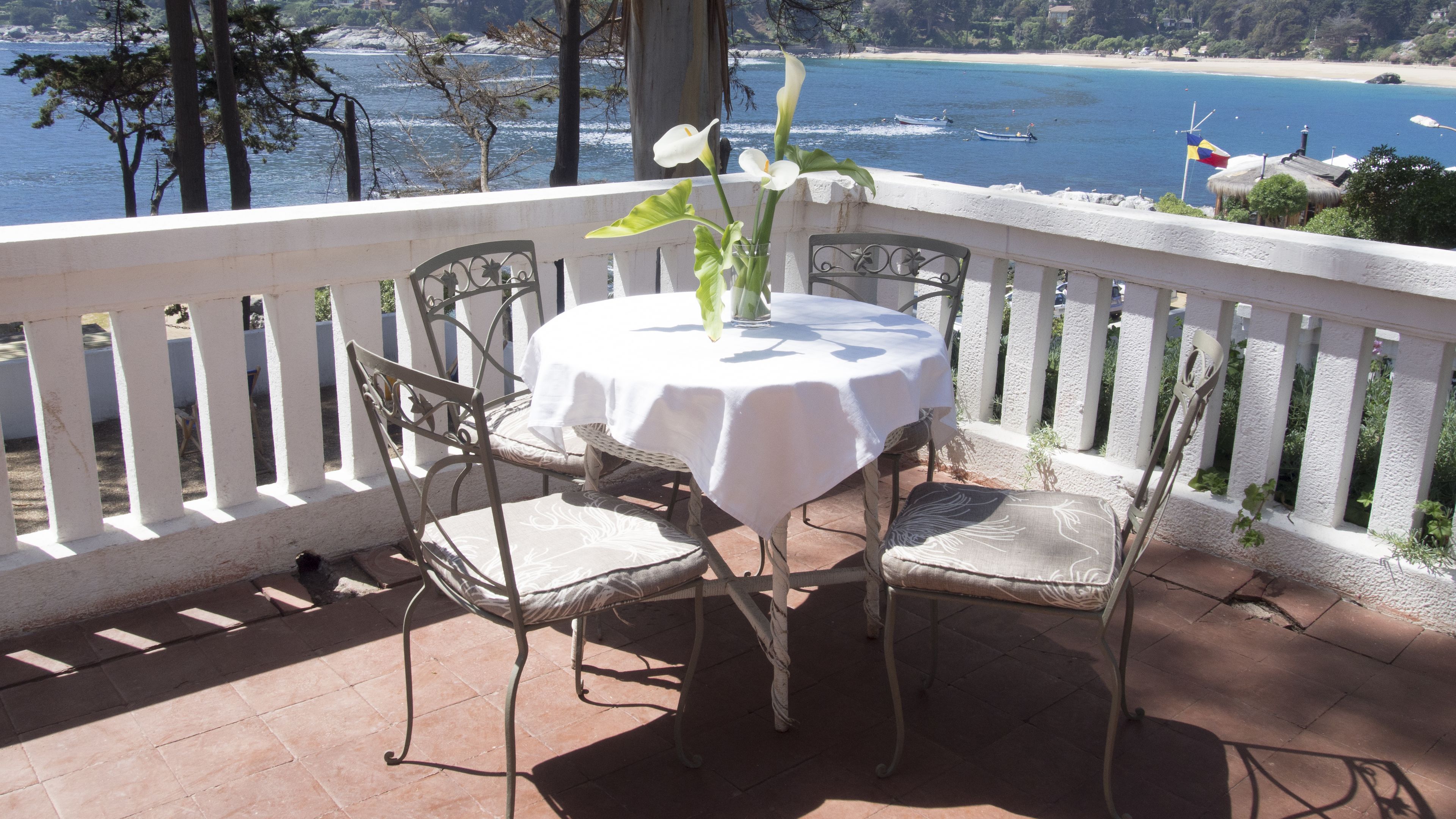 A patio with a table and chairs, overlooking a beach with boats and a flag.