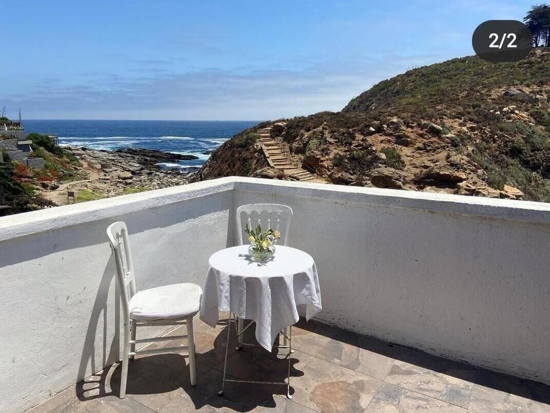 A balcony with a white table and chair, overlooking a rocky coastline and the ocean.
